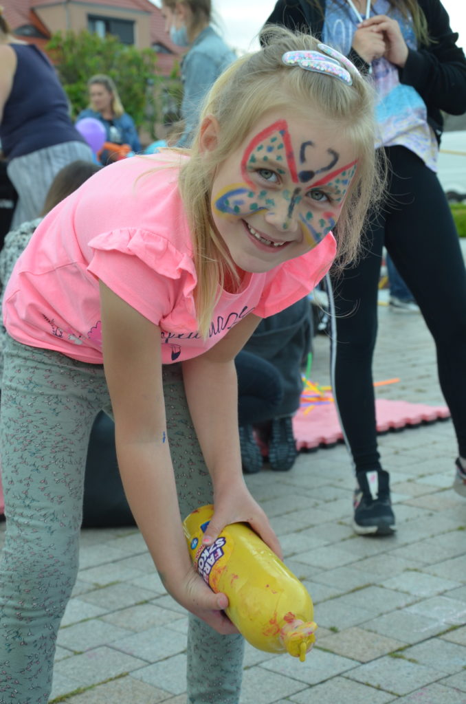 Na obrazku dziewczynka z kolorową twarzą trzyma butelkę z farbą