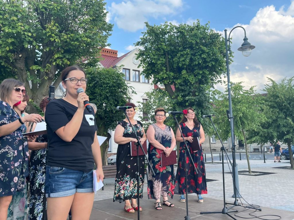 Na obrazku stoi kobieta z mikrofonem oraz kilka kobiet