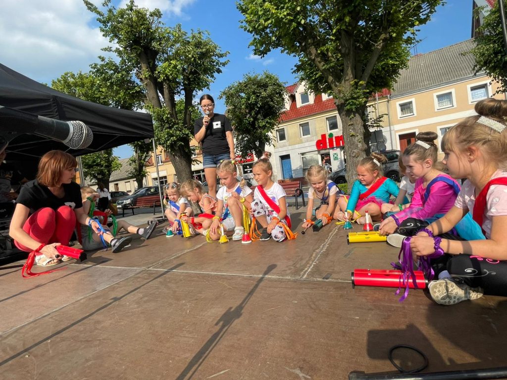 Na obrazku grupa dzieci siedzi z kobietą na scenie